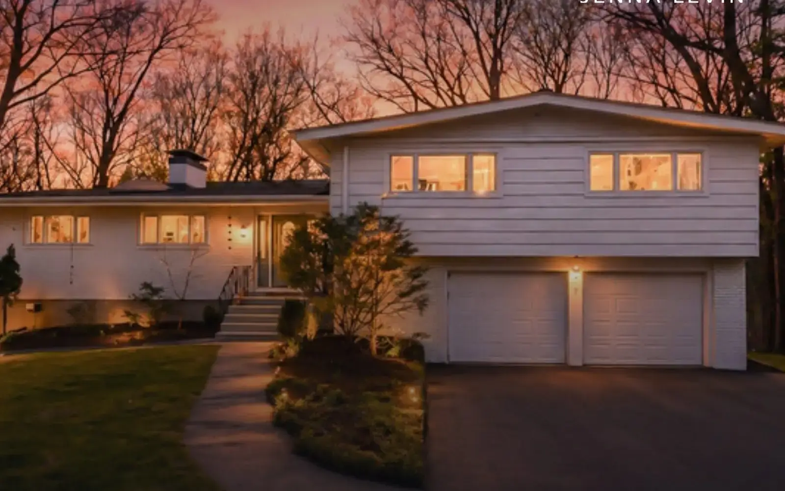 White split-level home with a double garage and glowing windows beneath a pink dusk sky.